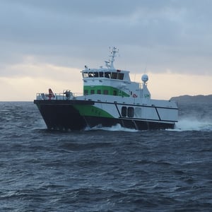 Nopea työalus, jonka runko on vihreä ja valkoinen ja joka kulkee karulla avomerellä pilvisen taivaan alla.