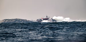 Nopea pelastusalus navigoi kovalla merellä aaltojen paukkuessa, taustalla näkyy kallioinen rantaviiva.